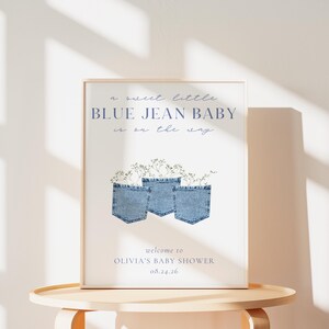 May include: A framed baby shower sign with the text "a sweet little BLUE JEAN BABY is on the way." The sign features three blue jean pockets with floral accents. The bottom of the sign reads "welcome to OLIVIA'S BABY SHOWER 08.24.26."