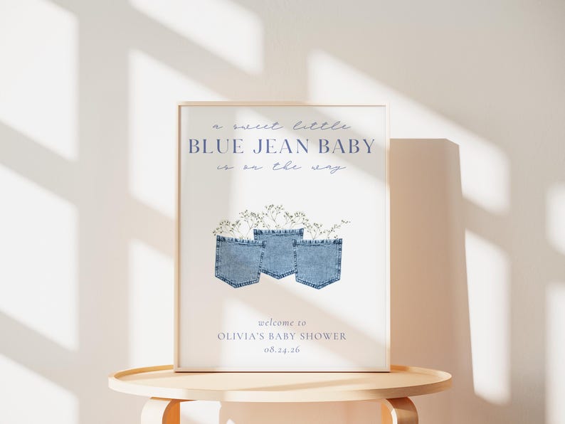 May include: A framed baby shower sign with the text "a sweet little BLUE JEAN BABY is on the way." The sign features three blue jean pockets with floral accents. The bottom of the sign reads "welcome to OLIVIA'S BABY SHOWER 08.24.26."