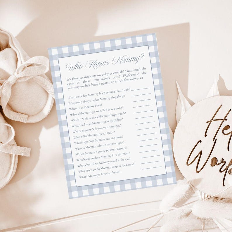 May include: A baby shower game card with a blue gingham border and the text "Who Knows Mommy?" in elegant script. The card includes fill-in-the-blank questions about the mother-to-be. A wooden sign that says "Her World" is also in the image.