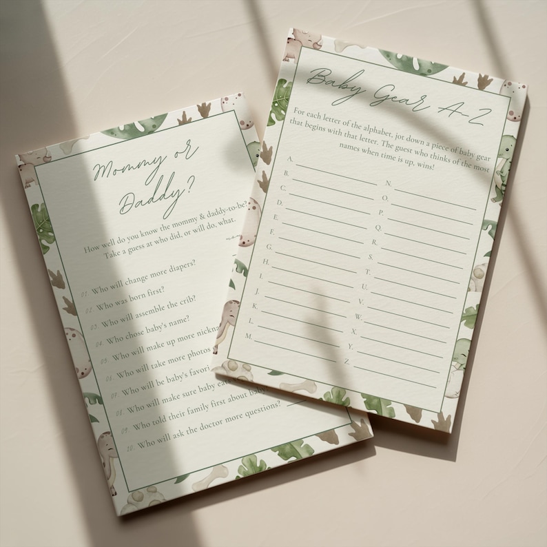 Puede incluir: Dos tarjetas de juego para baby shower. Una tarjeta se titula "&iquest;Mam&aacute; o Pap&aacute;?" con preguntas para completar. La otra tarjeta se titula "Baby Gear A-Z" con l&iacute;neas alfab&eacute;ticas. Ambas tarjetas tienen un fondo beige claro con un borde de hojas y animales verdes.