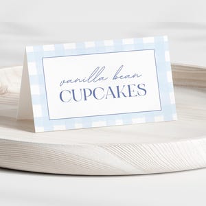 Könnte beinhalten: Eine weiße Karte mit den Worten "vanilla bean CUPCAKES" in blauer Schrift. Die Karte hat einen hellblauen Vichy-Rand und wird auf einem hellen Holztablett präsentiert.