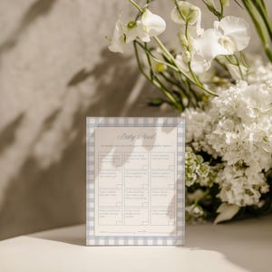 May include: A baby shower game card titled "Baby Feud" with a light blue gingham border. The card features fill-in-the-blank prompts for guests to guess popular baby-related answers. White flowers are in the background.