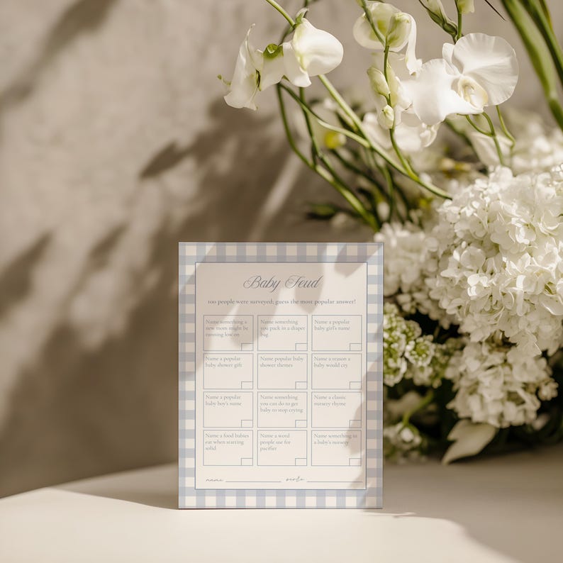 May include: A baby shower game card titled "Baby Feud" with a light blue gingham border. The card features fill-in-the-blank prompts for guests to guess popular baby-related answers. White flowers are in the background.