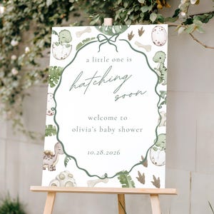 Puede incluir: Un letrero de baby shower sobre un caballete de madera. El letrero presenta un diseño verde y blanco con ilustraciones de dinosaurios y el texto "a little one is hatching soon". También se muestra la fecha "10.28.2026".