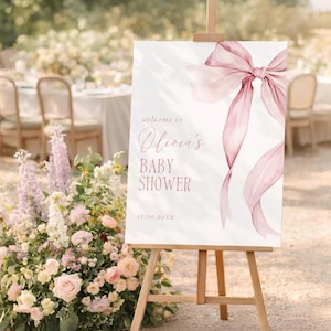 Könnte beinhalten: Eine Holzstaffelei zeigt ein weißes Schild mit der Aufschrift "welcome to Olivia's BABY SHOWER" in zartrosa Schrift. Eine große rosa Schleife schmückt die obere rechte Ecke. Das Schild ist von Blumen umgeben.