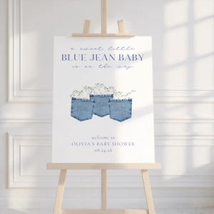 May include: A baby shower sign on a wooden easel. The sign features the text "a sweet little BLUE JEAN BABY is on the way" in blue script. Below are three denim jean pockets with floral accents. The sign reads "welcome to OLIVIA'S BABY SHOWER 08.24.26".