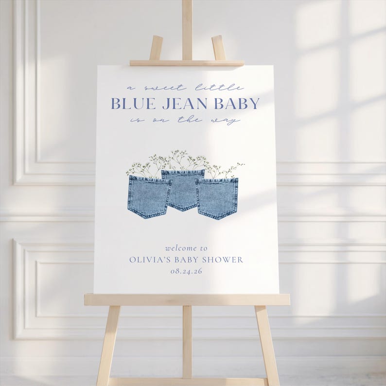 May include: A baby shower sign on a wooden easel. The sign features the text "a sweet little BLUE JEAN BABY is on the way" in blue script. Below are three denim jean pockets with floral accents. The sign reads "welcome to OLIVIA'S BABY SHOWER 08.24.26".