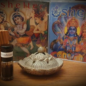 Può includere: Una composizione con due CD con la scritta "shelter", una piccola bottiglia con l'etichetta "Vrindavan", una scatola di legno e un bruciatore di incenso in ceramica a forma di loto. I CD presentano opere d'arte religiosa colorate.