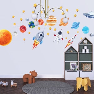 May include: A set of space-themed wall decals featuring planets, stars, a rocket, an astronaut, and a UFO. The decals are displayed on a light blue wall in a child's room.