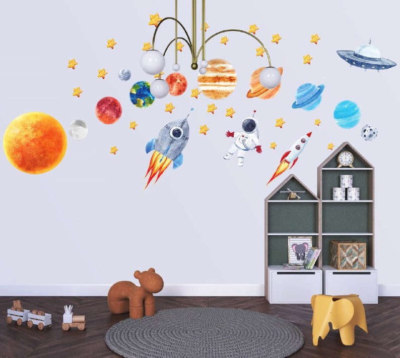 May include: A set of space-themed wall decals featuring planets, stars, a rocket, an astronaut, and a UFO. The decals are displayed on a light blue wall in a child's room.