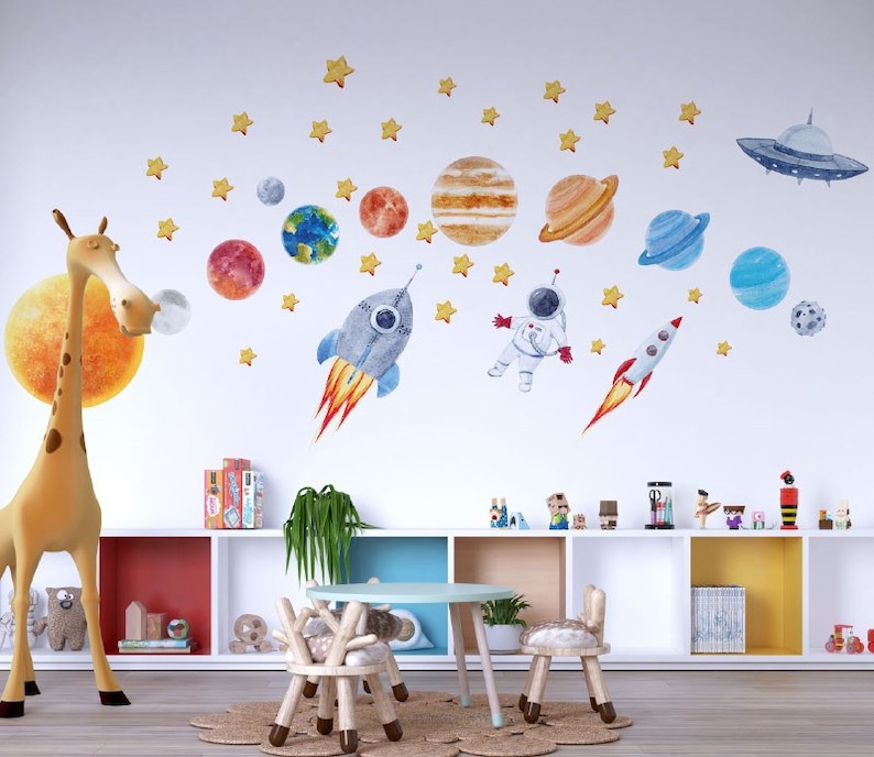 May include: A white wall with a space-themed wall decal featuring planets, stars, a rocket, an astronaut, and a flying saucer. A giraffe figurine stands in front of the wall. A bookshelf with toys and books is below the wall decal.