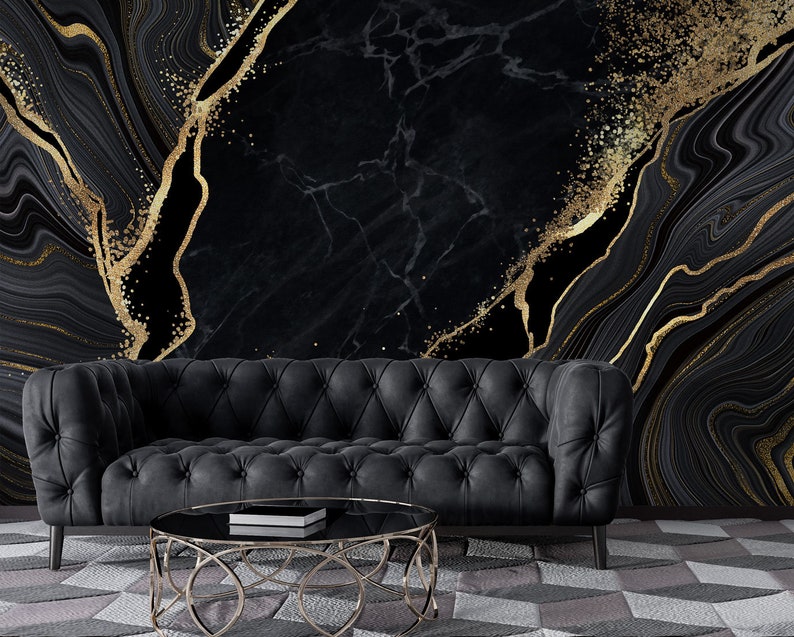 May include: A black tufted sofa with rolled arms sits before a wall featuring a black and gold marble-patterned mural. A round coffee table with a gold metal frame and a black glass top is positioned in front of the sofa on a geometric rug.