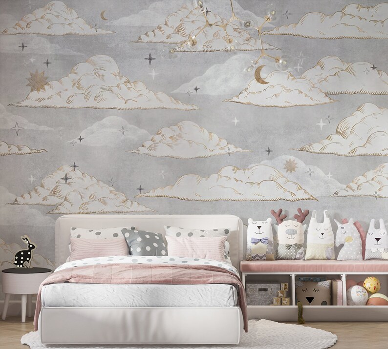May include: A bedroom featuring a white bed with pink and grey polka dot pillows, a pink throw blanket, and a white storage bench holding five animal-shaped plush toys. The wall is covered in grey wallpaper with gold-outlined clouds, stars, and moons.