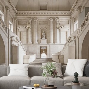 Op de afbeelding: Een ruime, lichte interieurruimte met een grote trap en sierlijke architectonische details. De sc&egrave;ne omvat een grijze hoekbank met witte kussens, een glazen vaas met groen en een kleine sculptuur op een ronde salontafel.