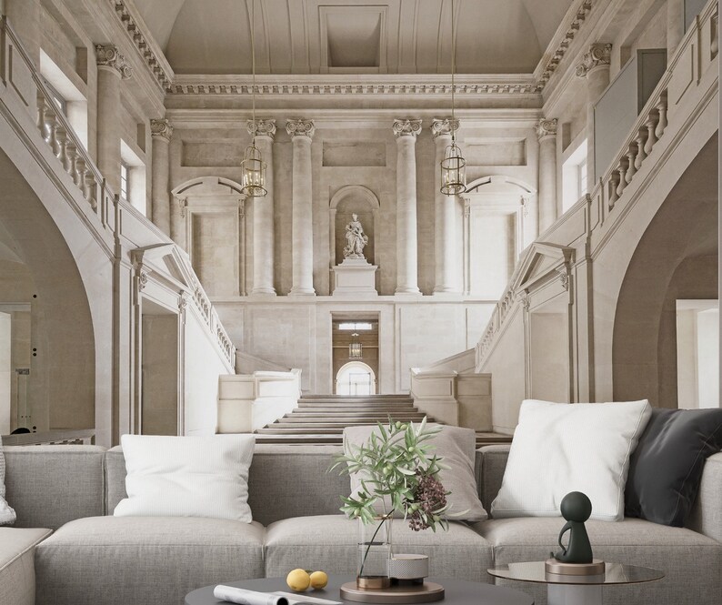 Op de afbeelding: Een ruime, lichte interieurruimte met een grote trap en sierlijke architectonische details. De sc&egrave;ne omvat een grijze hoekbank met witte kussens, een glazen vaas met groen en een kleine sculptuur op een ronde salontafel.