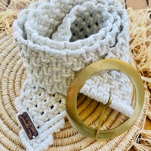 Peut inclure: Une ceinture en crochet blanc avec une boucle en laiton. La ceinture est enroulée et posée sur un tapis en paille tressée.