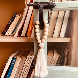 May include: A wooden bead garland with a white tassel hangs from a shelf with books. The books have titles in French and English. The shelf is made of wood and has a glass door.