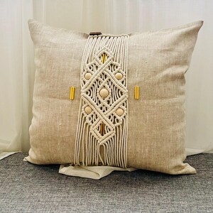May include: Beige linen pillow with a cream macrame design. The macrame features a geometric pattern with wooden beads and gold accents. The pillow is square and rests on a gray surface.