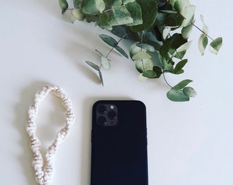 May include: A black phone case with a white phone strap. The phone case has a camera lens with four holes. The phone case is on a white surface with a bouquet of green eucalyptus leaves.