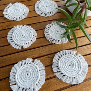 Peut inclure: Six dessous de verre en macramé blancs, avec un motif en spirale et des bords à franges, sont disposés sur une table en bois. Les dessous de verre sont ronds et semblent faits à la main. Une plante en pot avec des feuilles vertes est en arrière-plan.