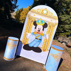 May include: A blue and gold backdrop with a cartoon image of Mickey Mouse dressed as a prince. Two blue and gold decorative columns are in front of the backdrop.