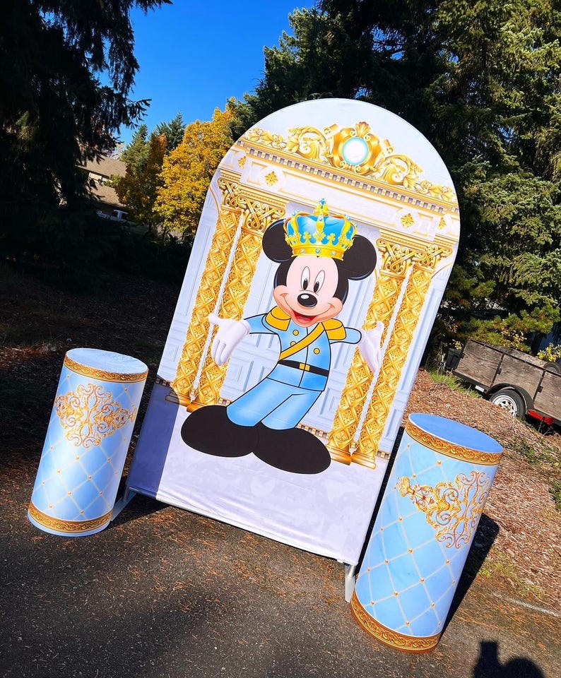 May include: A blue and gold backdrop with a cartoon image of Mickey Mouse dressed as a prince. Two blue and gold decorative columns are in front of the backdrop.