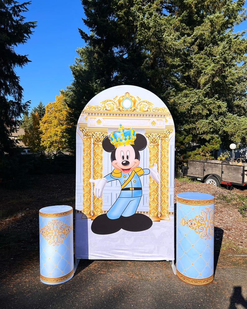 May include: A blue and gold themed party backdrop featuring Mickey Mouse dressed as a king. The backdrop is arched and has a gold trim. Two blue and gold columns are on either side of the backdrop.