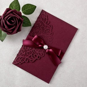 May include: Burgundy invitation envelope with a laser-cut design and a burgundy satin ribbon tied with a pearl embellishment.