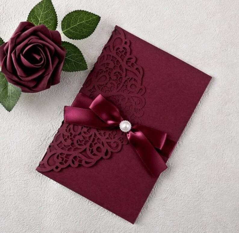May include: Burgundy invitation envelope with a laser-cut design and a burgundy satin ribbon tied with a pearl embellishment.