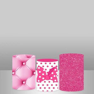 May include: Three pink cylindrical cake stands with different patterns. The first stand has a pink quilted design, the second stand has a white background with pink polka dots and a pink bow, and the third stand has a pink glitter design.