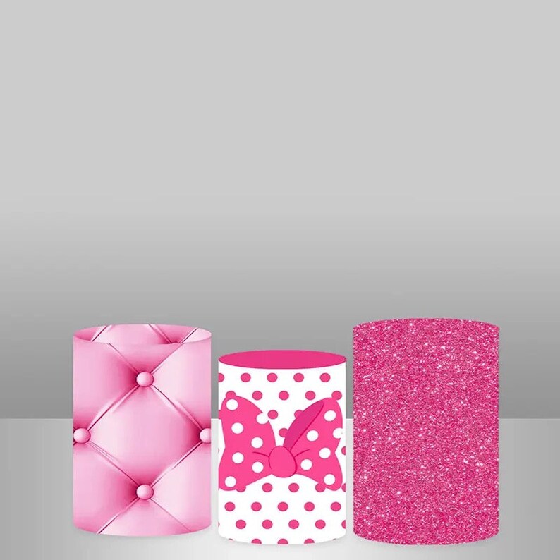 May include: Three pink cylindrical cake stands with different patterns. The first stand has a pink quilted design, the second stand has a white background with pink polka dots and a pink bow, and the third stand has a pink glitter design.