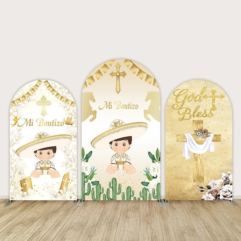 May include: Three white arch-shaped photo booth backdrops with gold accents. The center backdrop features a boy wearing a sombrero and the text "Mi Bautizo". The left backdrop features a similar boy and the text "Mi Bautizo". The right backdrop features a cross with the text "God Bless".