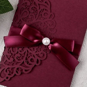 May include: Burgundy wedding invitation with a laser cut design and a burgundy satin ribbon with a pearl embellishment.