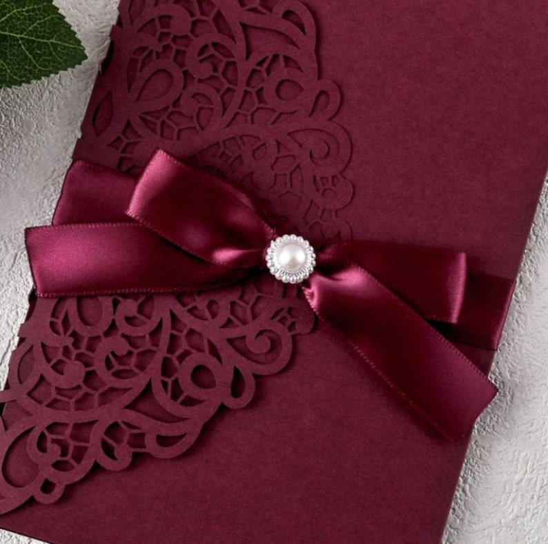 May include: Burgundy wedding invitation with a laser cut design and a burgundy satin ribbon with a pearl embellishment.