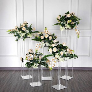 May include: Five clear acrylic pedestal stands of varying heights, each topped with a white floral arrangement. The flowers include roses, orchids, and greenery.