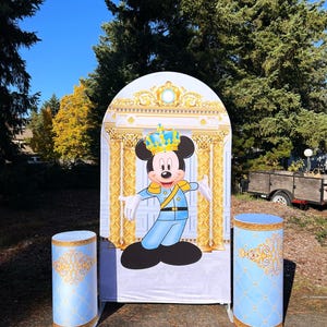 May include: A blue and gold themed party backdrop featuring Mickey Mouse dressed as a king. The backdrop is arched and has a gold trim. Two blue and gold pillars are in front of the backdrop.