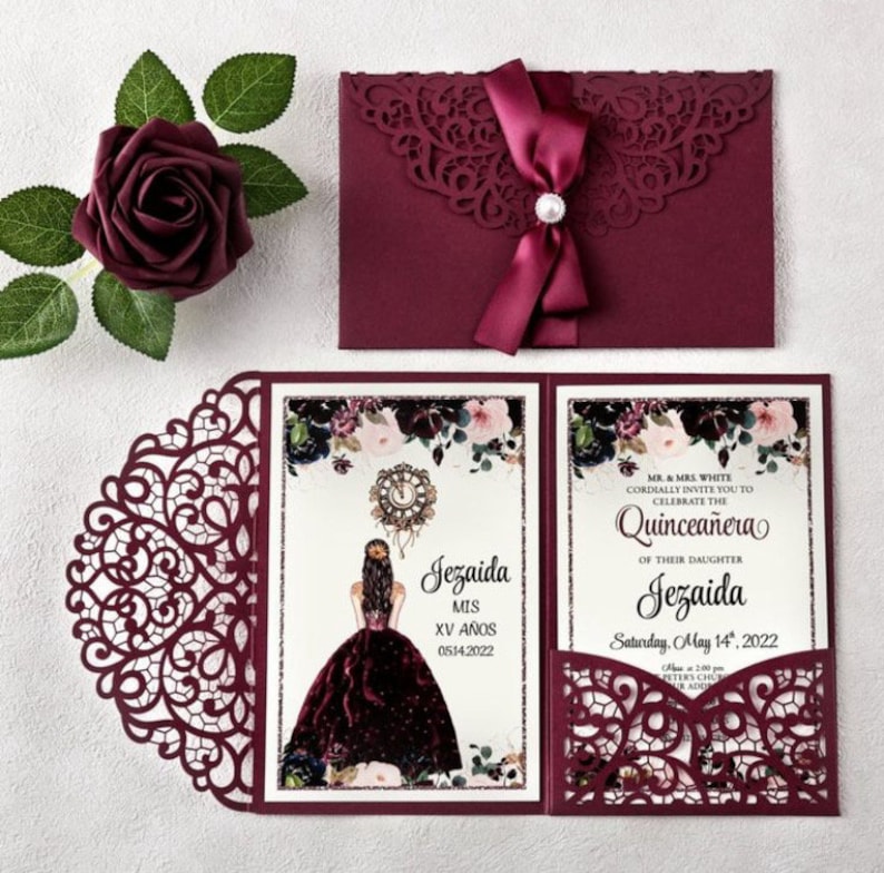May include: Burgundy Quincea&ntilde;era invitation with a lace design. The invitation features a pocket for a RSVP card and a burgundy ribbon with a pearl accent. The text on the invitation reads "Jezaida MIS XV A&Ntilde;OS 0514.2022" and "MR. & MRS. WHITE CORDIALLY INVITE YOU TO CELEBRATE THE QUINCEA&Ntilde;ERA OF THEIR DAUGHTER Jezaida Saturday, May 14, 2022".