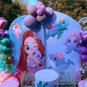 May include: A mermaid themed party backdrop with a purple, green, and pink balloon arch. The backdrop features a mermaid with a purple octopus, a turtle, and a dolphin. There are two white pillars with blue and green designs.