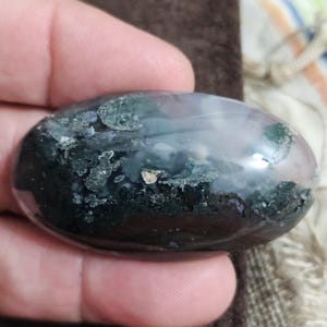 May include: A polished, oval gemstone displaying a blend of dark green, black, and pale purple tones. The stone has a smooth, glossy finish and is held in a hand. The gemstone's colours and patterns create a natural, organic aesthetic.