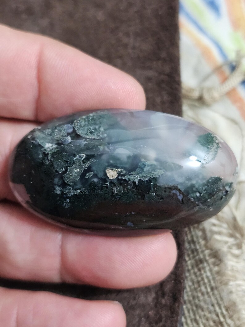 May include: A polished, oval gemstone displaying a blend of dark green, black, and pale purple tones. The stone has a smooth, glossy finish and is held in a hand. The gemstone's colours and patterns create a natural, organic aesthetic.