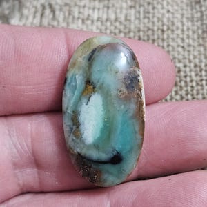 May include: An oval-shaped gemstone with a polished surface. The stone features a blend of colors, including teal, brown, black, and white. The gemstone is held in a hand, showcasing its natural beauty and unique color patterns.