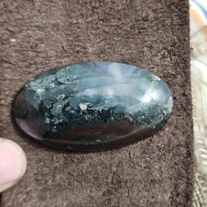 May include: An oval gemstone with a polished surface. The stone has a mix of dark green, black, and translucent areas, creating a unique pattern. The gemstone is set against a brown, textured background.