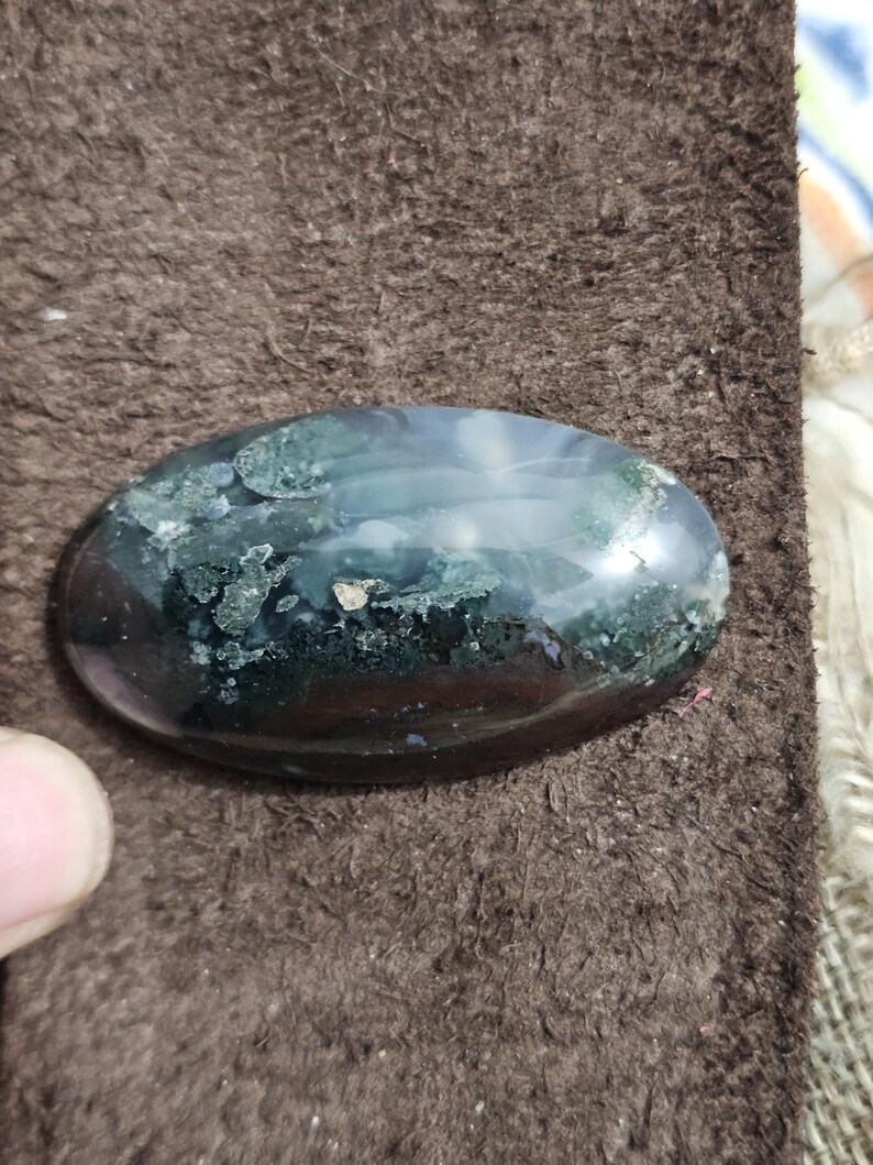 May include: An oval gemstone with a polished surface. The stone has a mix of dark green, black, and translucent areas, creating a unique pattern. The gemstone is set against a brown, textured background.