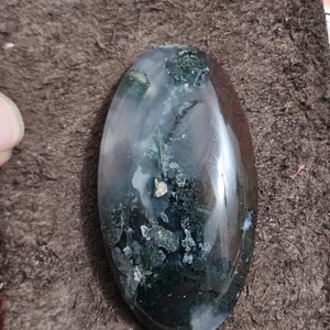 May include: An oval gemstone with a polished surface. The stone displays a mix of clear, white, green, and brown colours. The green areas have a mossy appearance. The gemstone is set against a brown background.