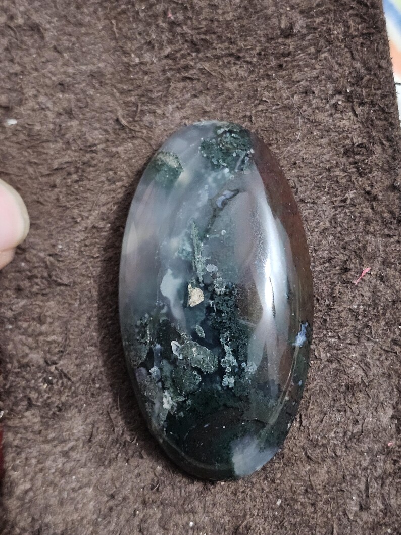 May include: An oval gemstone with a polished surface. The stone displays a mix of clear, white, green, and brown colours. The green areas have a mossy appearance. The gemstone is set against a brown background.