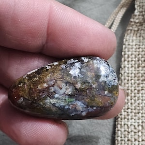 May include: A polished, oval-shaped stone with a mottled pattern of brown, white, and green hues. The stone's surface has a glossy finish, and it is held in a person's hand. The stone is a natural product.