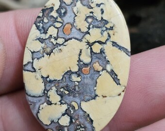 May include: Oval-shaped gemstone with a unique pattern. The stone features a cream-colored base with a network of gray, black, and orange-brown inclusions. The polished surface highlights the natural variations, making it suitable for jewelry or crafts.
