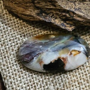 May include: A close-up of a colorful, oval-shaped agate stone with brown, white, and black swirls. The stone is sitting on a woven fabric background.
