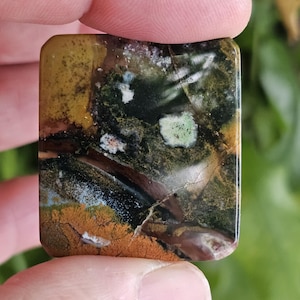 Può includere: Una gemma rettangolare con una superficie lucida. La pietra presenta un mix di colori, tra cui marrone, verde e giallo. Il motivo include aree di verde scuro, verde chiaro e marrone-arancio. La pietra è tenuta tra due dita.