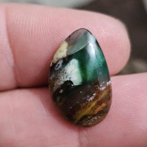 May include: A polished, teardrop-shaped gemstone with a mix of colors. The stone features areas of deep green, brown, and white, with hints of yellow and gray. The surface is smooth and reflective, showcasing natural patterns.